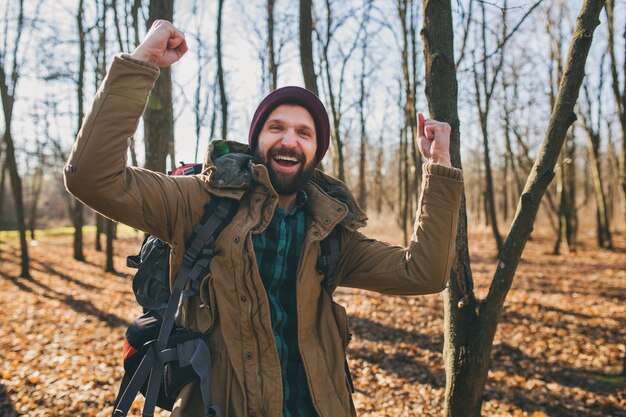 Hombre joven inconformista viajando con mochila en el bosque de otoño con chaqueta y sombrero, turista activo, explorando la naturaleza en la estación fría