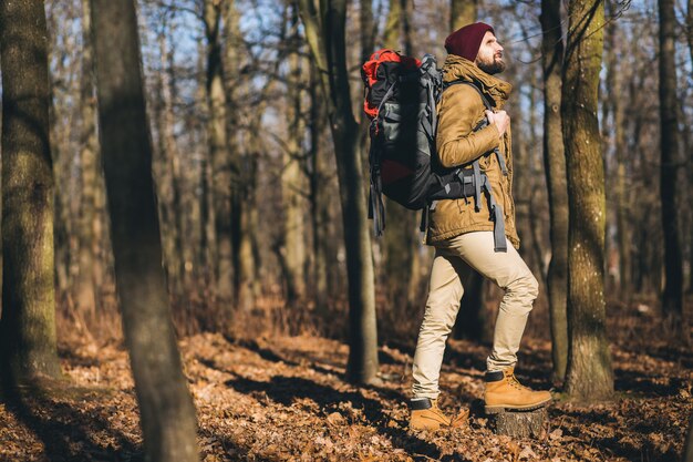 Hombre joven inconformista viajando con mochila en el bosque de otoño con chaqueta y sombrero, turista activo, explorando la naturaleza en la estación fría