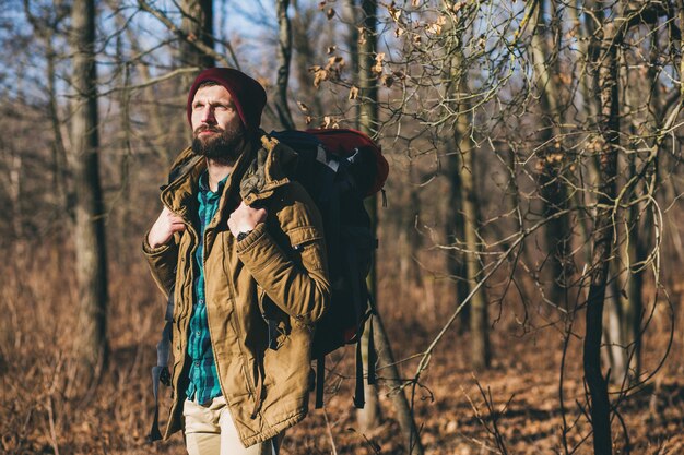 Hombre joven inconformista viajando con mochila en el bosque de otoño con chaqueta y sombrero, turista activo, explorando la naturaleza en la estación fría