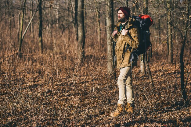 Hombre joven inconformista viajando con mochila en el bosque de otoño con chaqueta y sombrero, turista activo, explorando la naturaleza en la estación fría