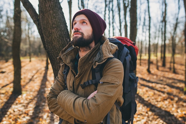 Hombre joven inconformista viajando con mochila en el bosque de otoño vistiendo chaqueta y sombrero