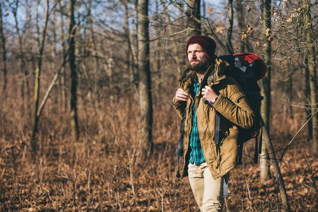 Hombre joven inconformista viajando con mochila en el bosque de otoño vistiendo chaqueta y sombrero