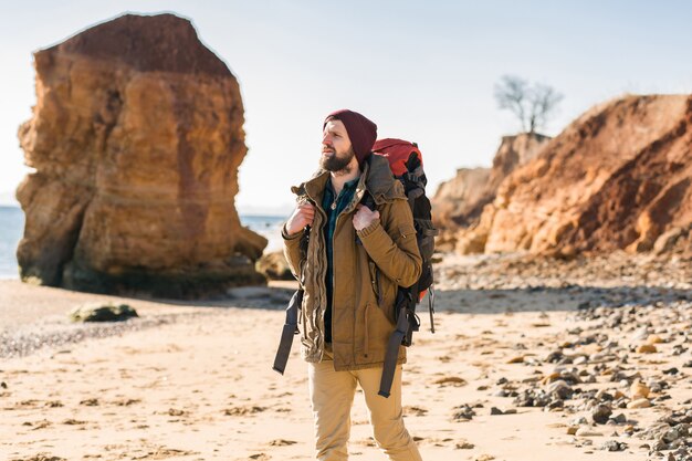 Hombre joven inconformista viajando con mochila en la costa del mar de otoño vistiendo chaqueta y sombrero