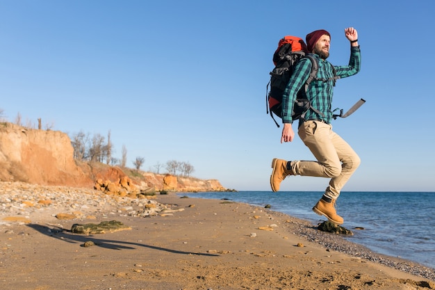 Hombre joven inconformista viajando con mochila en la costa del mar de otoño vistiendo chaqueta y sombrero