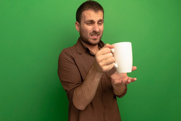 Hombre joven irritado aislado en verde sosteniendo y mirando una taza de té manteniendo la mano en la pared de aire