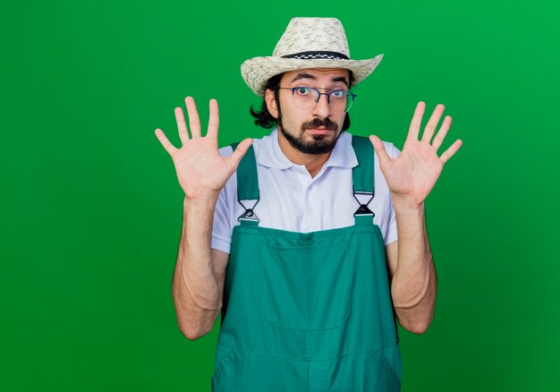 Hombre joven jardinero barbudo vestido con mono y sombrero levantando las palmas mirando confundido sin respuesta