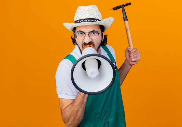 Hombre joven jardinero barbudo vestido con mono y sombrero sosteniendo mini rastrillo gritando al megáfono con expresión agresiva de pie sobre la pared naranja