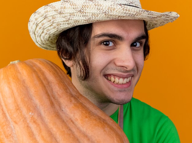 Foto gratuita hombre joven jardinero complacido en delantal y sombrero mostrando calabaza sonriendo ampliamente con cara feliz de pie sobre fondo naranja