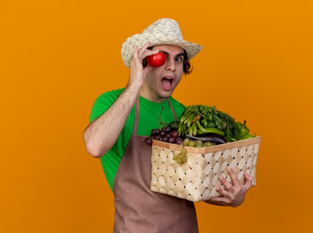 Hombre joven jardinero en delantal y sombrero con caja llena de verduras y tomate fresco cerca de su ojo sonriendo mirando sorprendido de pie sobre fondo naranja