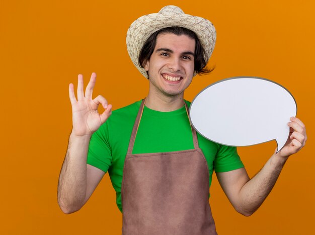 Hombre joven jardinero en delantal y sombrero con cartel de burbujas de discurso en blanco mirando a la cámara sonriendo ampliamente mostrando signo de ok de pie sobre fondo naranja