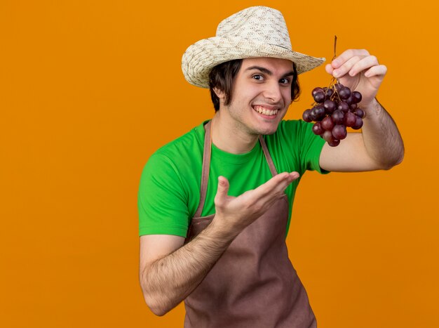 Hombre joven jardinero en delantal y sombrero mostrando racimo de uva que presenta con el brazo de la mano sonriendo de pie sobre la pared naranja