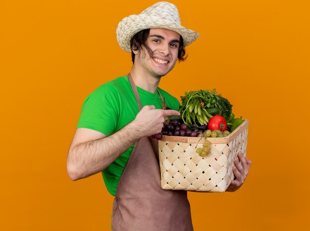 Hombre joven jardinero en delantal y sombrero sosteniendo la caja llena de verduras mirando a la cámara sonriendo alegremente apuntando con el dedo índice a la caja de pie sobre fondo naranja