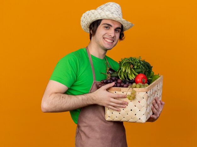 Hombre joven jardinero en delantal y sombrero sosteniendo la caja llena de verduras mirando a la cámara sonriendo con cara feliz de pie sobre fondo naranja