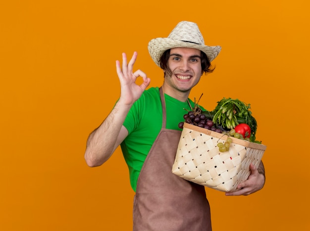 Hombre joven jardinero en delantal y sombrero sosteniendo una caja llena de verduras mirando sonriendo alegremente mostrando el signo de ok de pie sobre la pared naranja