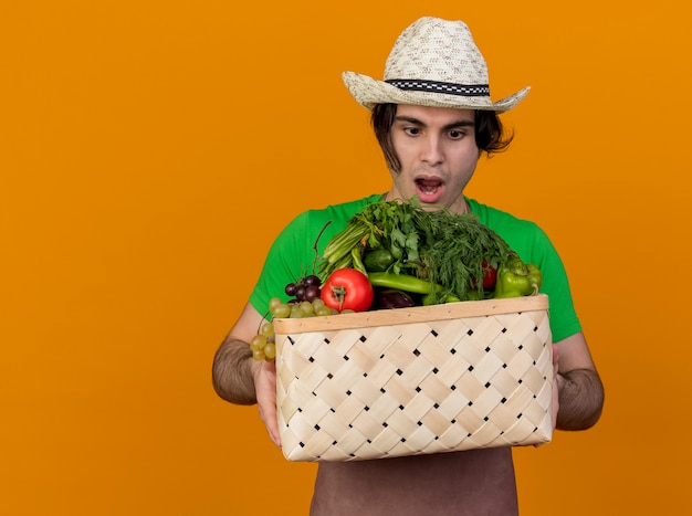 Hombre joven jardinero en delantal y sombrero sosteniendo cajón lleno de verduras mirando sorprendido y asombrado de pie sobre fondo naranja