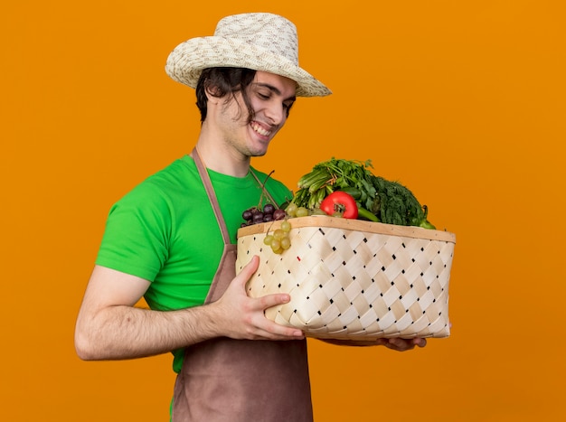 Foto gratuita hombre joven jardinero en delantal y sombrero sosteniendo cajón lleno de verduras mirándolo sonriendo con cara feliz de pie sobre la pared naranja