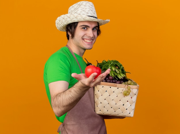 Hombre joven jardinero en delantal y sombrero sosteniendo cajón lleno de verduras mostrando tomate fresco sonriendo con cara feliz de pie sobre la pared naranja