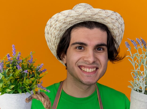 Hombre joven jardinero en delantal y sombrero sosteniendo plantas en macetas mirando sonriendo ampliamente con cara feliz de pie sobre la pared naranja