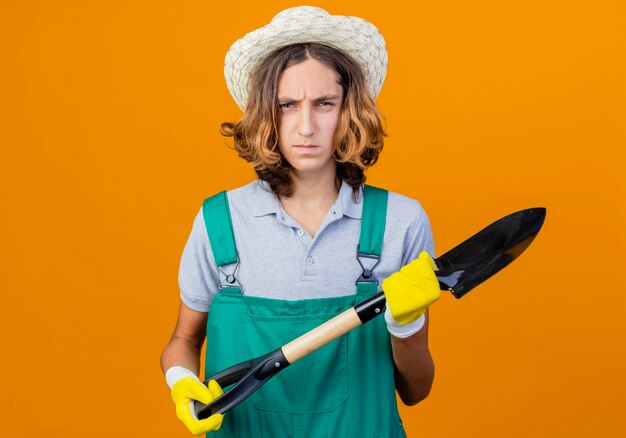 Hombre joven jardinero en guantes de goma con mono y sombrero con pala