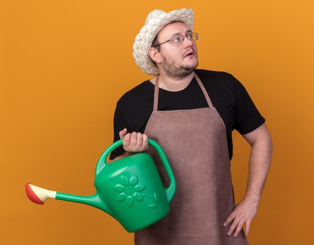 Hombre joven jardinero de lado mirando sorprendido con sombrero de jardinería sosteniendo regadera poniendo la mano en la cadera aislada en la pared naranja