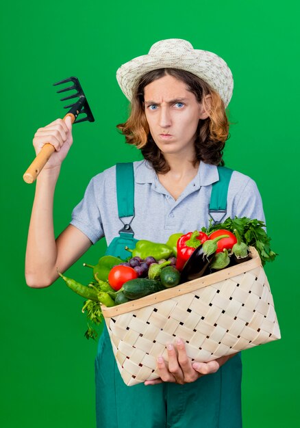 Hombre joven jardinero vestido con mono y sombrero con caja llena de verduras frescas