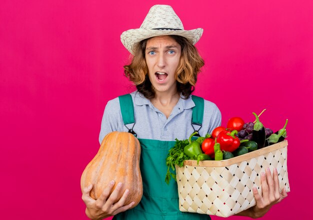 Hombre joven jardinero vestido con mono y sombrero con caja llena de verduras frescas