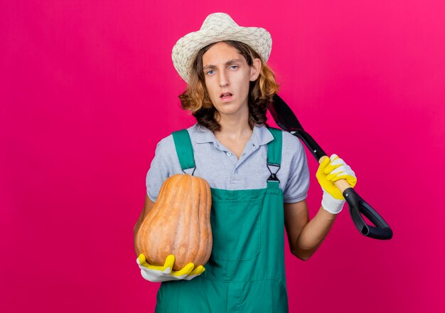 Hombre joven jardinero vestido con mono y sombrero con guantes de goma con pala