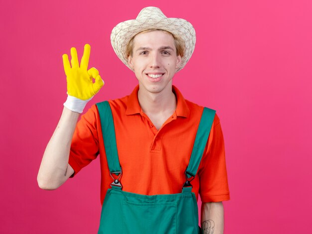 Hombre joven jardinero vestido con mono y sombrero en guantes de goma sonriendo mostrando signo ok