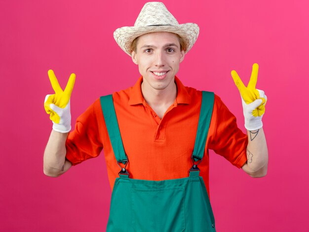 Hombre joven jardinero vestido con mono y sombrero en guantes de goma sonriendo mostrando signo v
