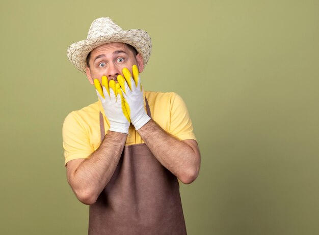 Hombre joven jardinero vestido con mono y sombrero en guantes de trabajo conmocionado cubriendo la boca con las manos