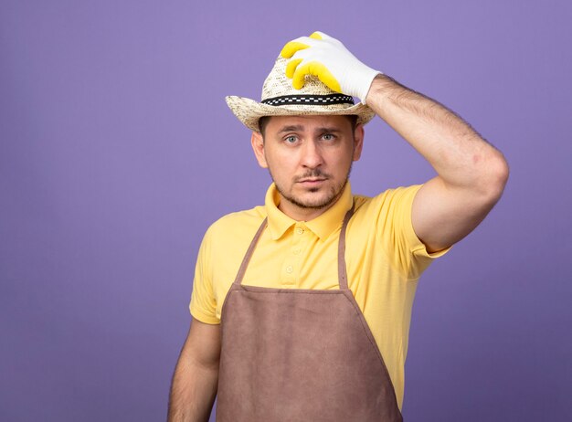 Hombre joven jardinero vestido con mono y sombrero en guantes de trabajo lookign confiado tocando su sombrero