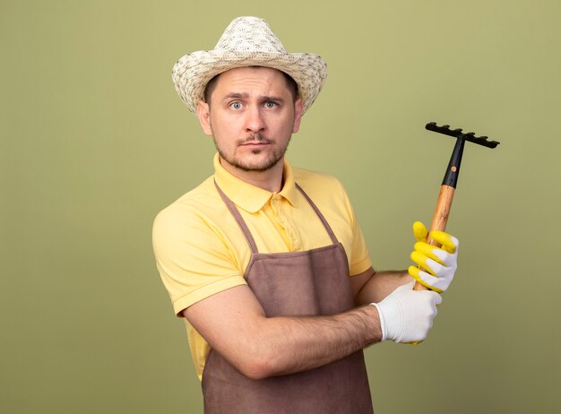 Hombre joven jardinero vestido con mono y sombrero en guantes de trabajo con mini rastrillo lookign a la cámara con cara seria