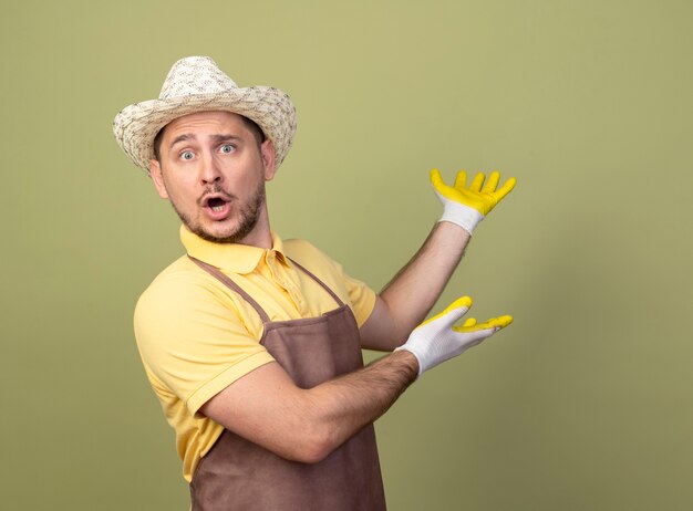 Hombre joven jardinero vestido con mono y sombrero en guantes de trabajo presentando algo con los brazos de las manos sorprendidos