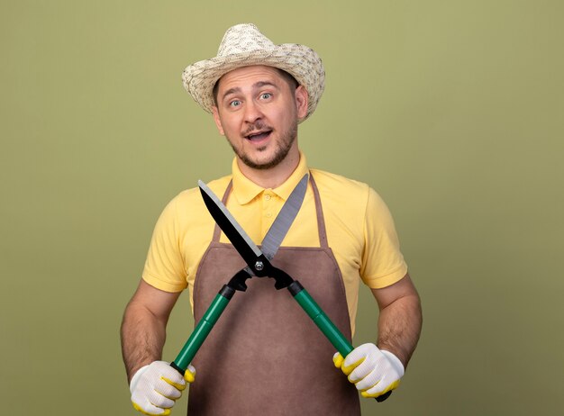 Hombre joven jardinero vestido con mono y sombrero en guantes de trabajo sosteniendo cortasetos feliz y sorprendido