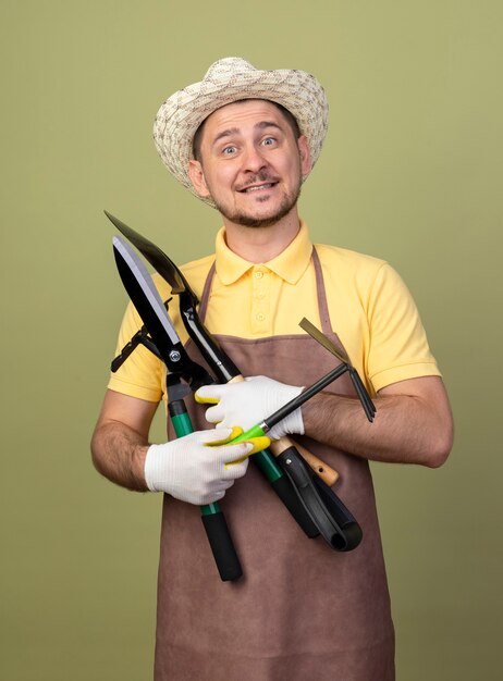 Hombre joven jardinero vestido con mono y sombrero en guantes de trabajo sosteniendo equipos de jardinería con una sonrisa en la cara