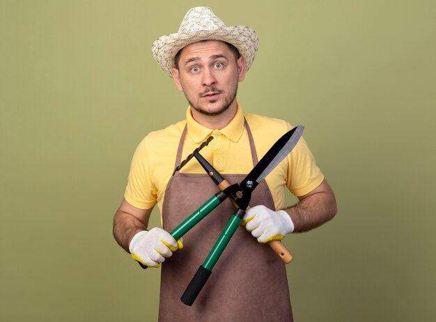 Hombre joven jardinero vestido con mono y sombrero en guantes de trabajo sosteniendo equipos de jardinería sorprendido