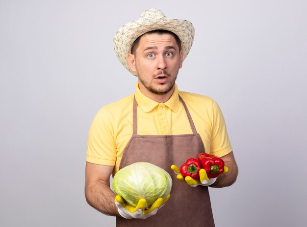 Hombre joven jardinero vestido con mono y sombrero en guantes de trabajo sosteniendo pimientos rojos y repollo confundido