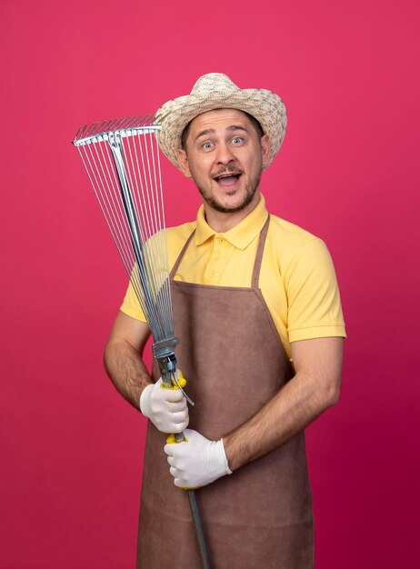 Hombre joven jardinero vestido con mono y sombrero en guantes de trabajo sosteniendo rastrillo sonriendo alegremente feliz y positivo