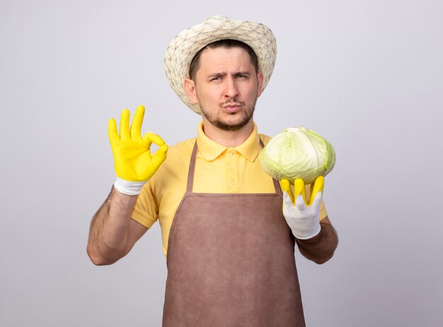 Hombre joven jardinero vestido con mono y sombrero en guantes de trabajo sosteniendo repollo mirando al frente con expresión de confianza que muestra el signo de ok de pie sobre la pared blanca