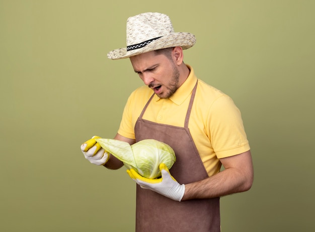 Hombre joven jardinero vestido con mono y sombrero en guantes de trabajo sosteniendo repollo mirándolo intrigado