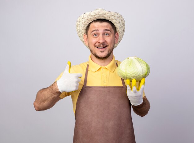 Hombre joven jardinero vestido con mono y sombrero en guantes de trabajo sosteniendo repollo sonriendo con cara feliz