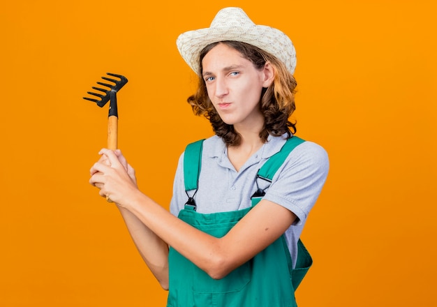 Hombre joven jardinero vestido con mono y sombrero mini rastrillo oscilante