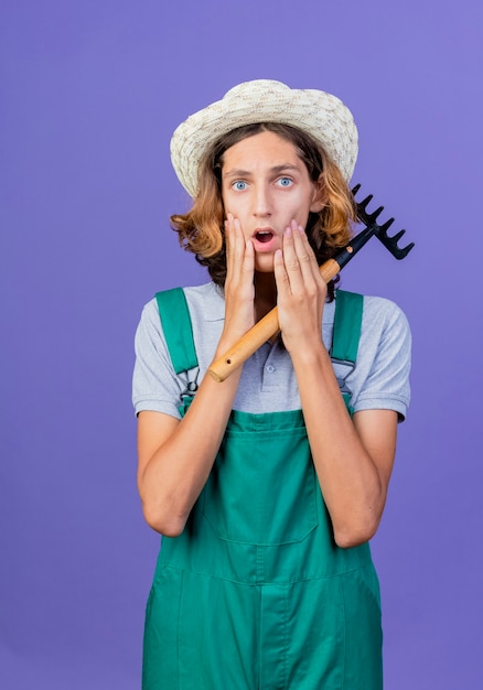 Hombre joven jardinero vestido con mono y sombrero con mini rastrillo