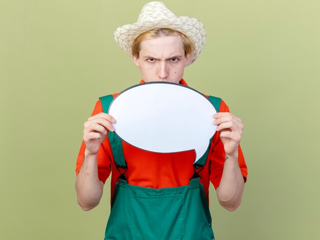 Hombre joven jardinero vestido con mono y sombrero que muestra el signo de la burbuja del discurso en blanco mirando a la cámara con la cara seria frunciendo el ceño sobre fondo claro