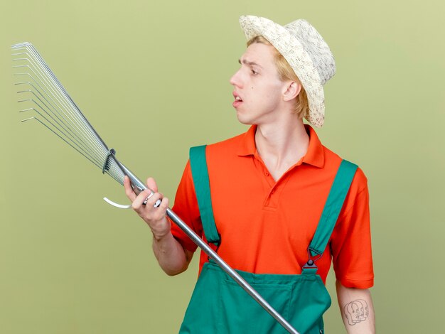 Foto gratuita hombre joven jardinero vestido con mono y sombrero con rastrillo