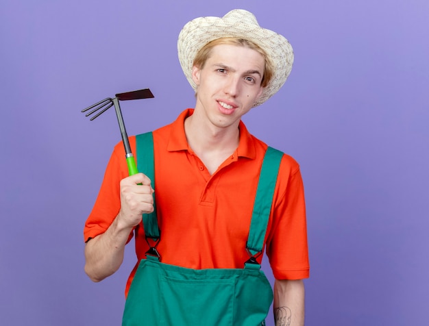 Hombre joven jardinero vestido con mono y sombrero sosteniendo azadón sonriendo confiado