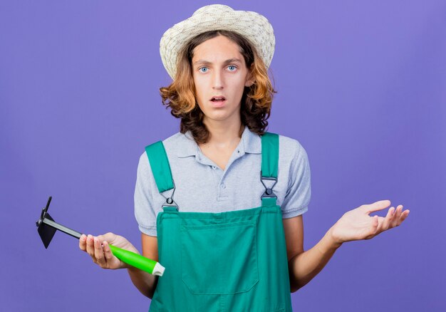 Hombre joven jardinero vestido con mono y sombrero sosteniendo azadón