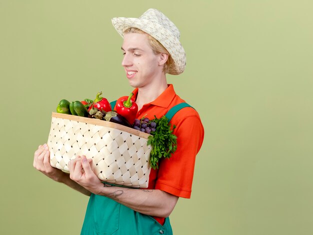 Hombre joven jardinero vestido con mono y sombrero sosteniendo la caja llena de verduras mirando verduras con una sonrisa en la cara de pie sobre fondo claro