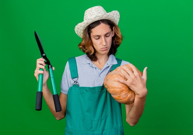 Hombre joven jardinero vestido con mono y sombrero sosteniendo calabaza y cortasetos