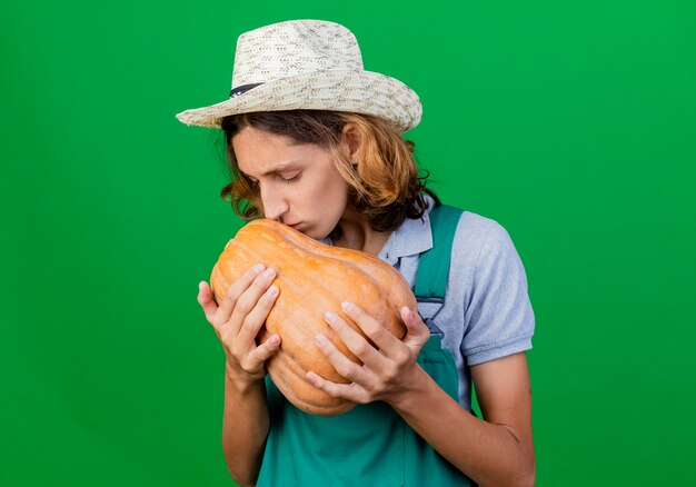 Hombre joven jardinero vestido con mono y sombrero sosteniendo calabaza en manos besándola